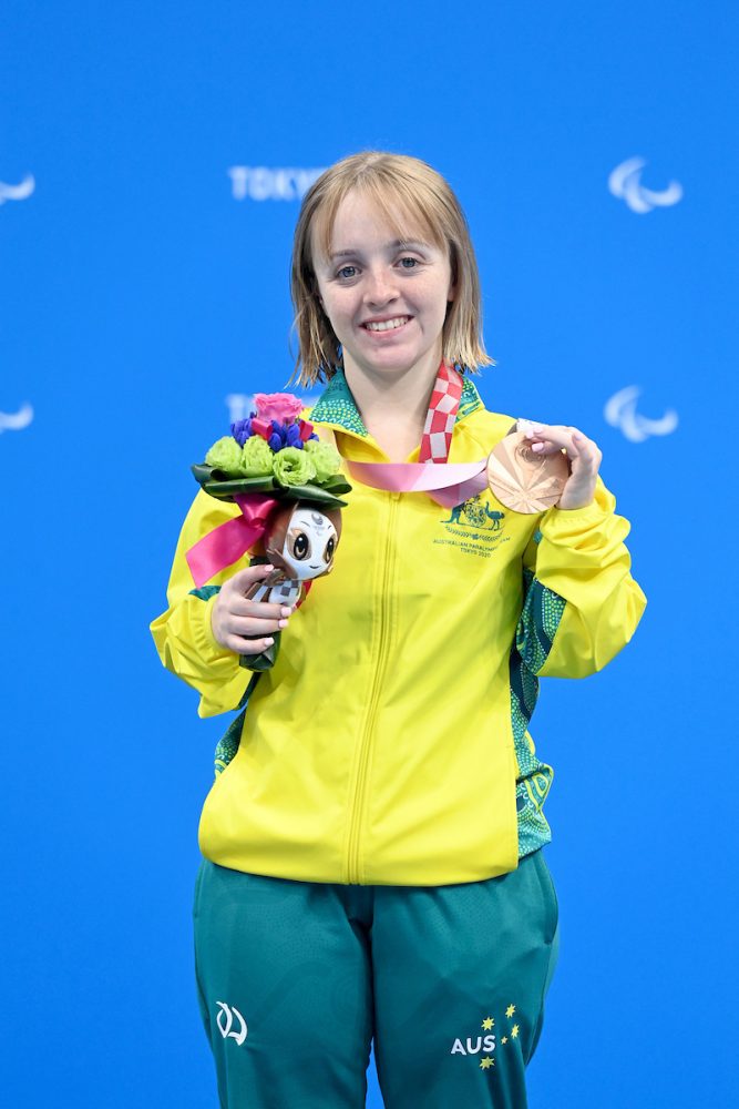 Para-swimmer Tiffany Thomas Kane holding her bronze medal. Wearing Green and Gold Australian Para-Athlete atire. Blue Backdrop.