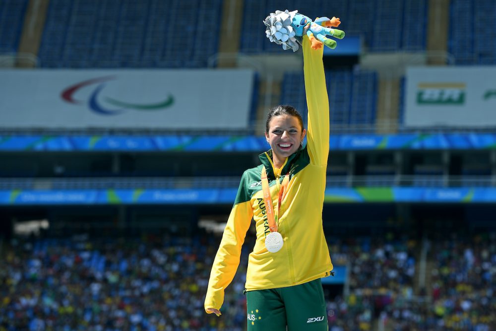 Para-athlete Taylor Doyle wearing silver medal and holding up Rio 2016 Mascot Plushie upwards in celebration. Dressed in green and gold para-athlete uniform. Stadium in the background.