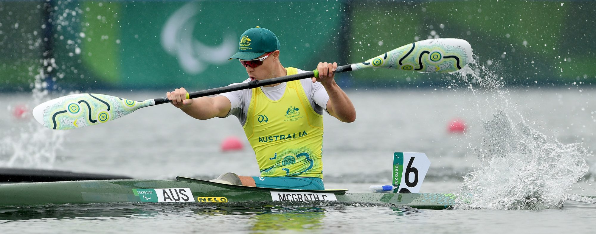 A male Australian Para-canoe athlete paddling. He is wearing a gold singlet and a green cap.